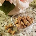 Bone-shaped baked dog treats made with apple and cinnamon, displayed in a glass container next to a flower vase on a kitchen counter