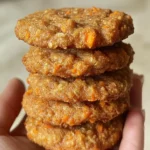 A hand holding a stack of homemade carrot and apple crunchy cookies for dogs, showing their golden texture and visible oat and carrot bits.